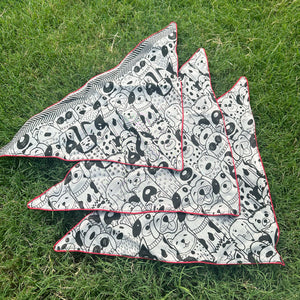 Black and white illustrated dog print cotton bandana with red border, photographed on green grass, by The Thread Weavers