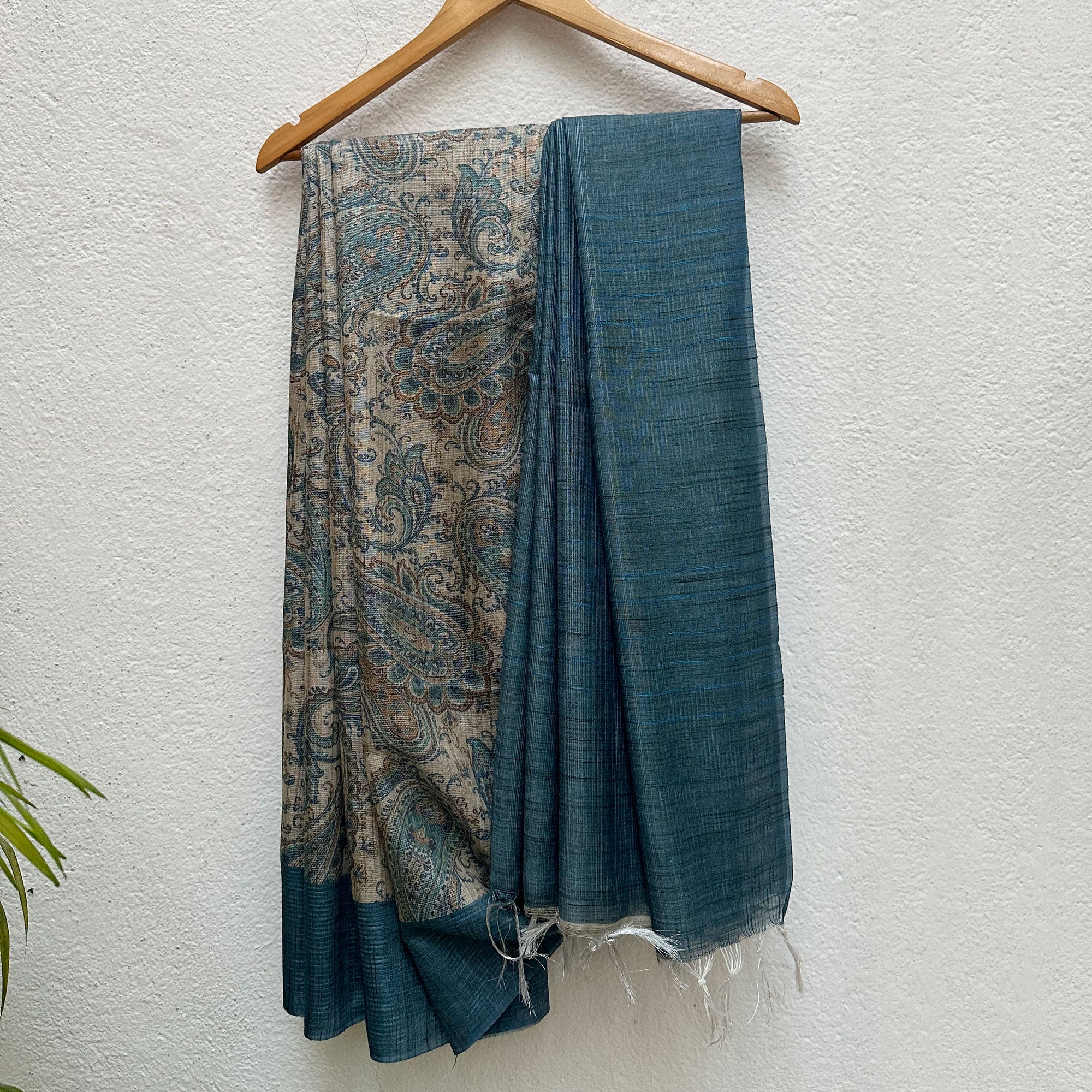 Ambi Printed Tissue Silk Saree in Tussar and teal blue with a bold paisley print, on a wooden hanger