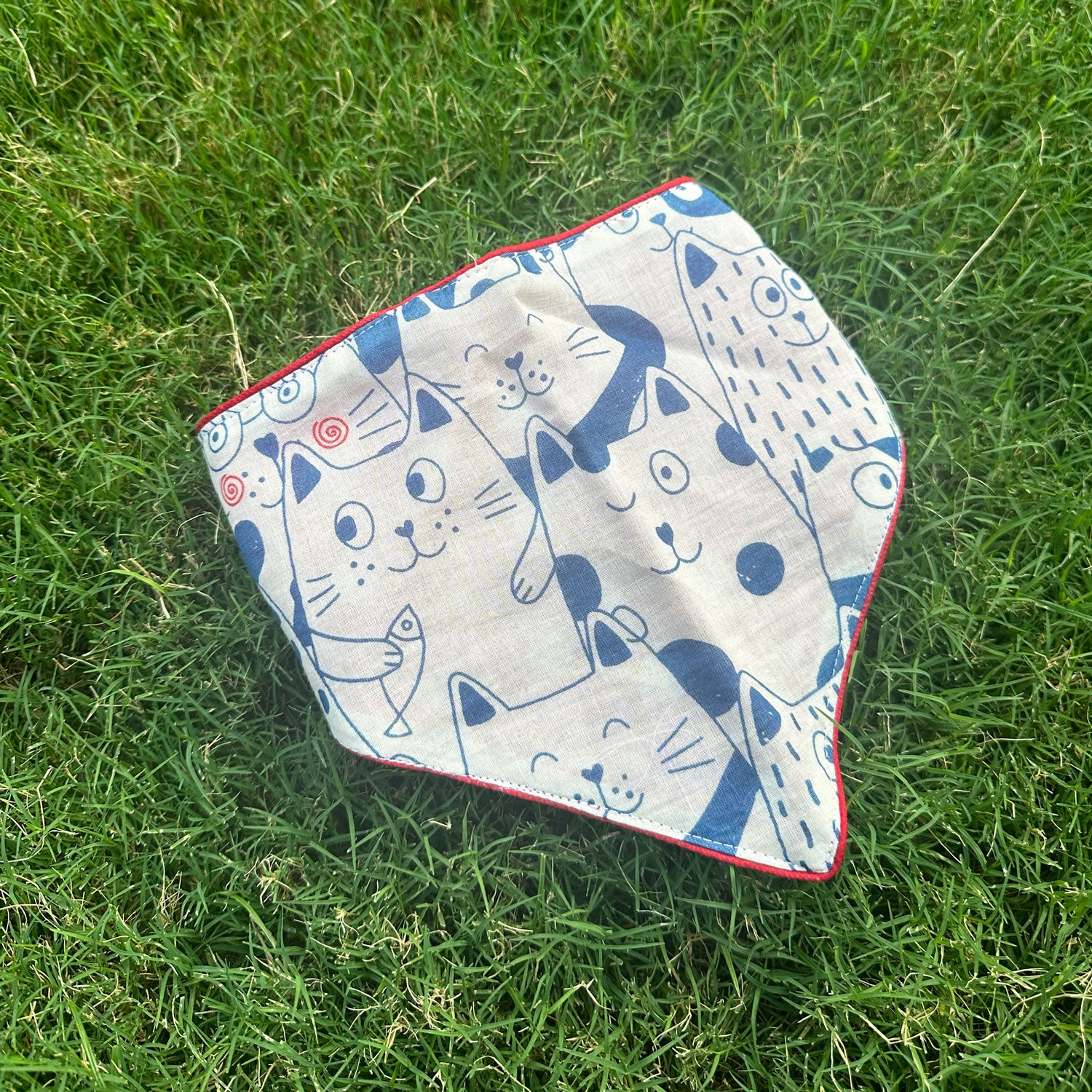 Navy and cream illustrated cat print cotton bandana with red border detail, photographed on green grass, by The Thread Weavers