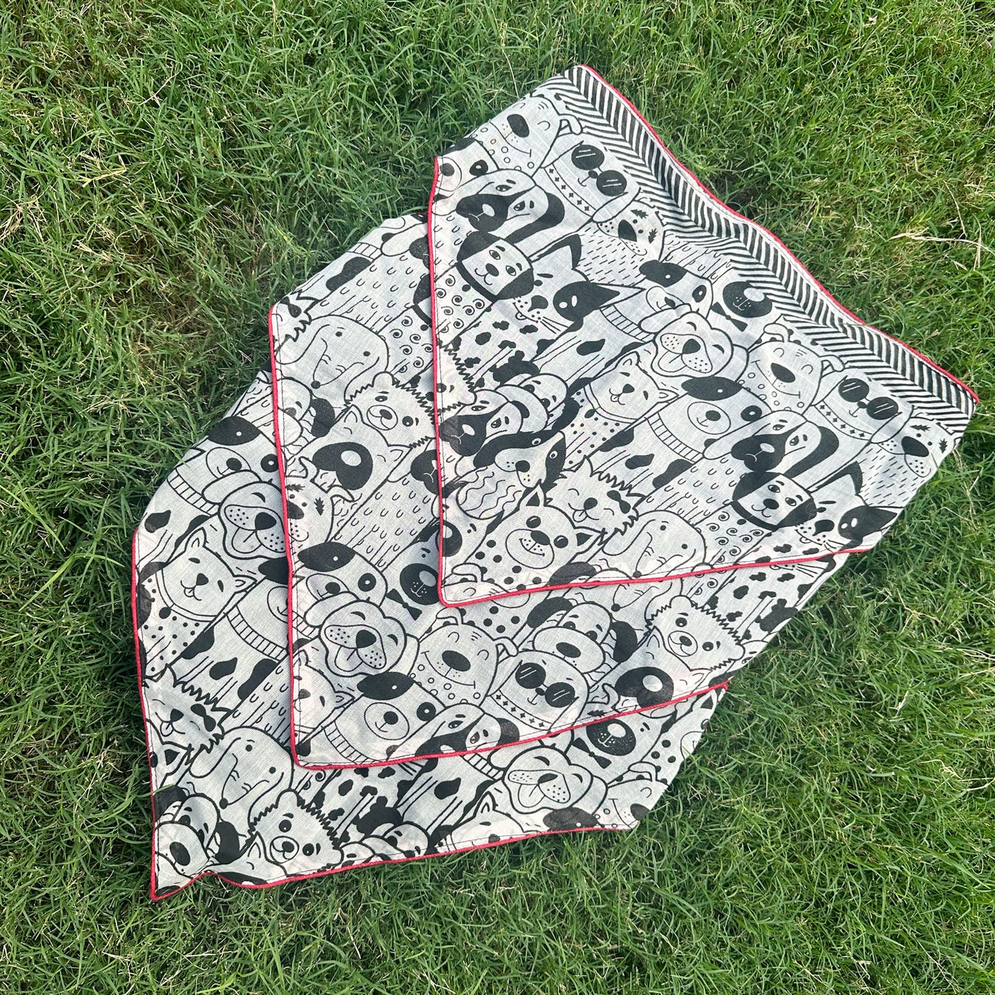 Black and white illustrated dog print cotton bandana with red border, photographed on green grass, by The Thread Weavers