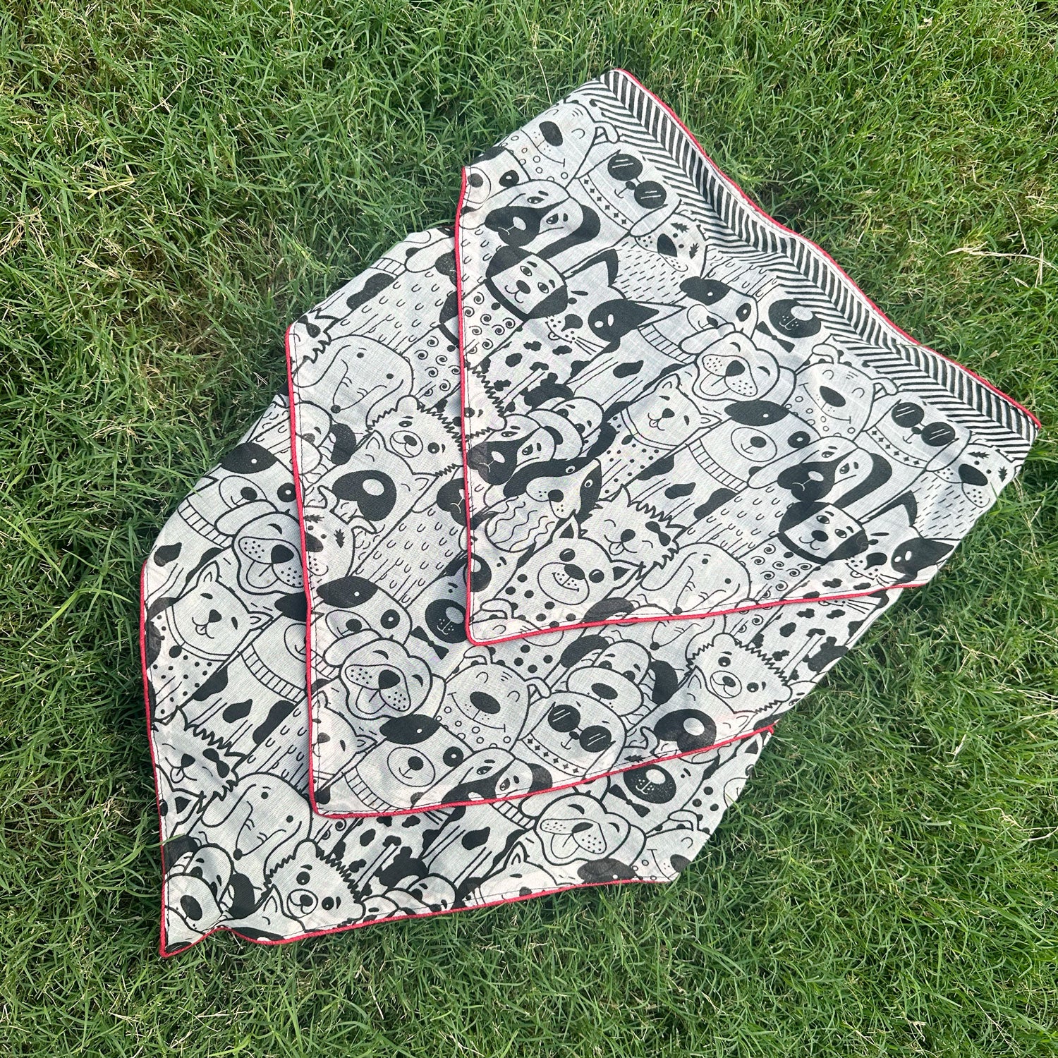 Black and white illustrated dog print cotton bandana with red border, photographed on green grass, by The Thread Weavers