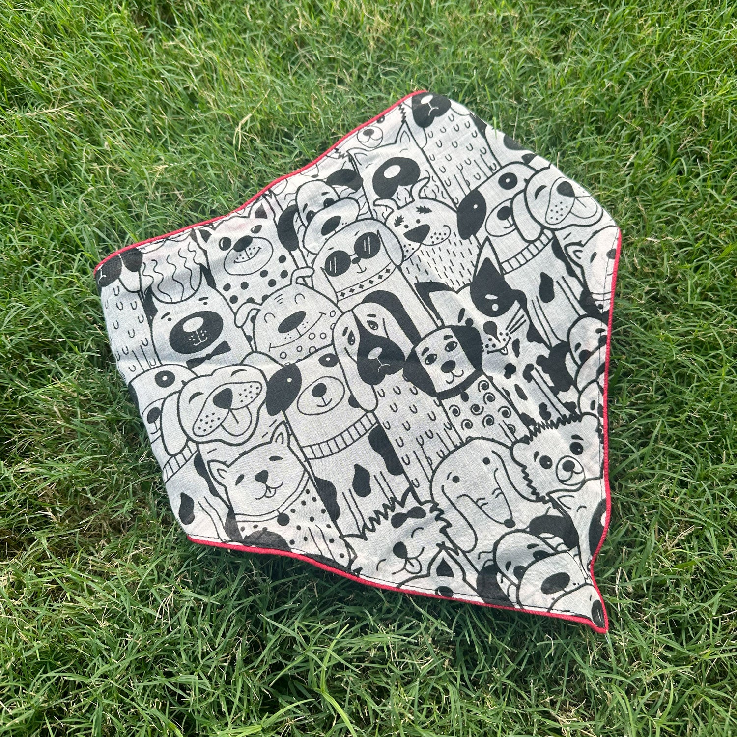 Black and white illustrated dog print cotton bandana with red border, photographed on green grass, by The Thread Weavers