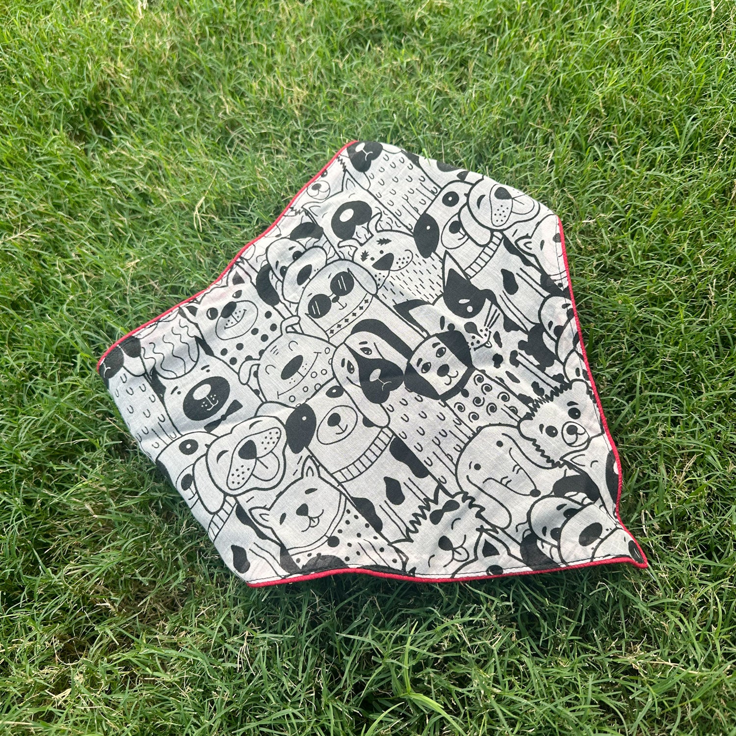 Black and white illustrated dog print cotton bandana with red border, photographed on green grass, by The Thread Weavers