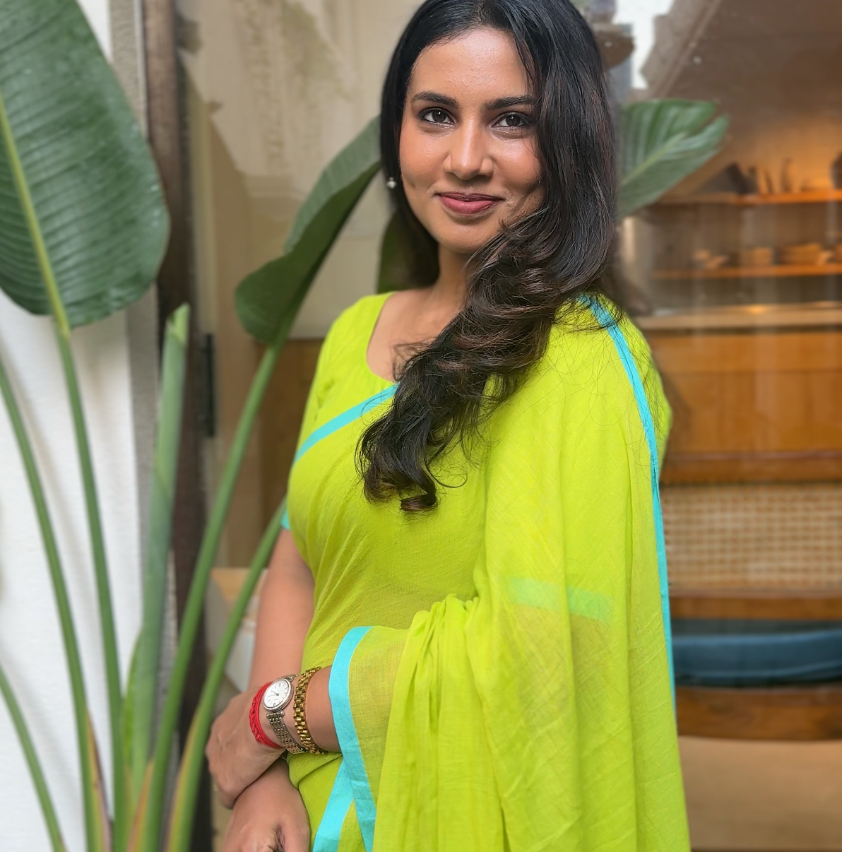 Woman in a bright green mul saree with blue border standing indoors with plants around.