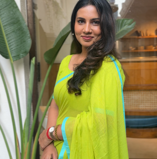 Woman in a bright green mul saree with blue border standing indoors with plants around.