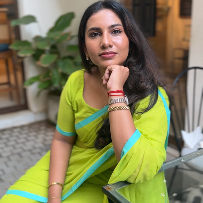 Woman in a bright green mul saree with blue borders sitting indoors. Perfect for Summers