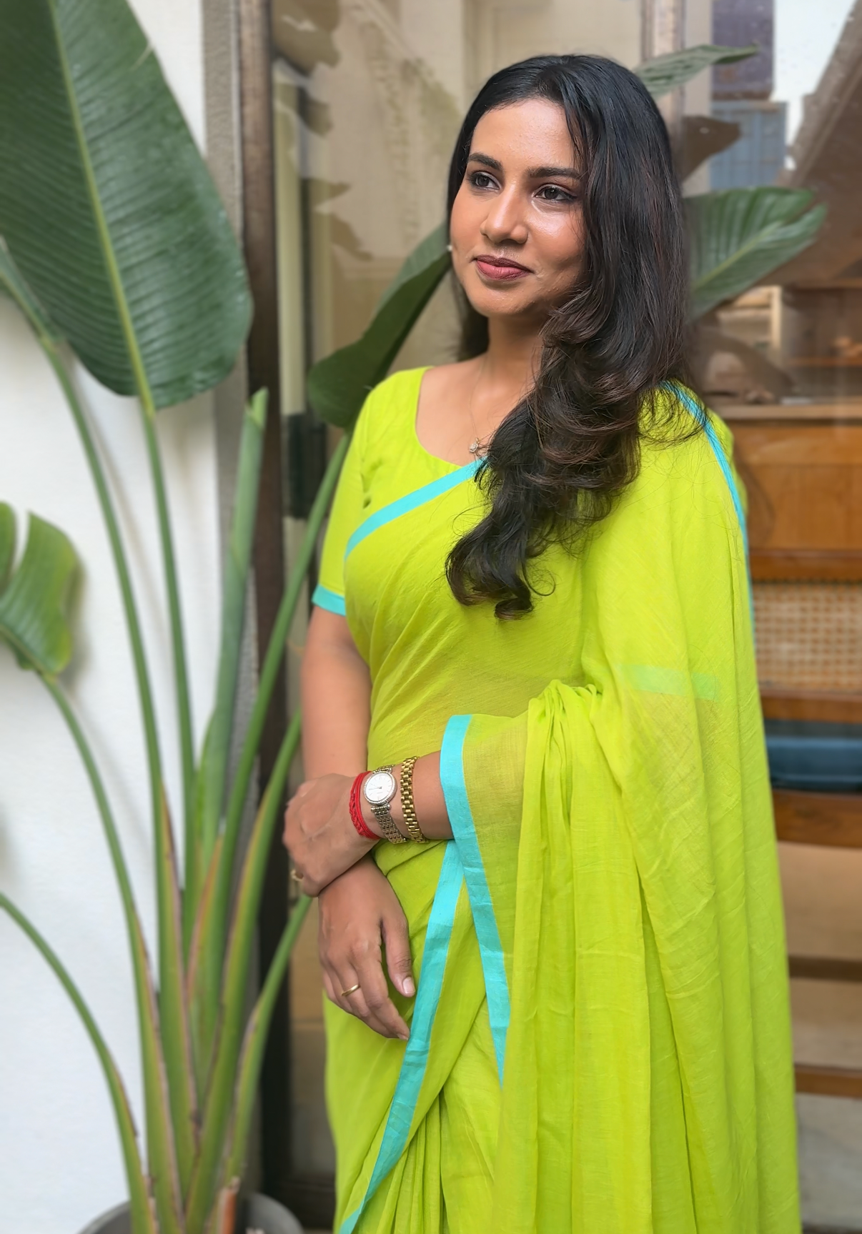Woman in a bright green saree with blue accents standing indoors next to a plant.