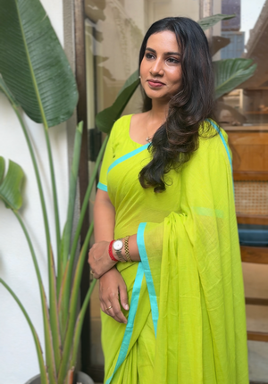 Woman in a bright green saree with blue accents standing indoors next to a plant.