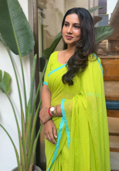 Woman in a bright green saree with blue accents standing indoors next to a plant.