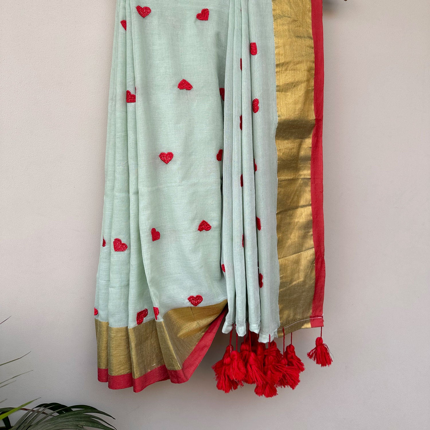 Contemporary mint green mul saree featuring delicate heart embroidery and contrast red tassel detailing.