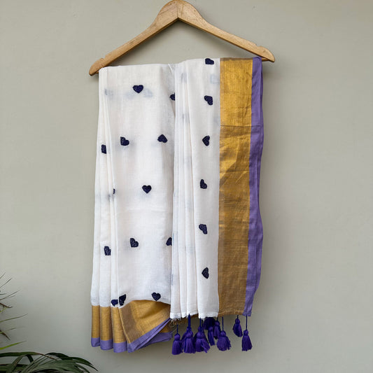 Moonmilk white mul cotton saree with lavender embroidered heart motifs, gold border and handcrafted purple tassels.
